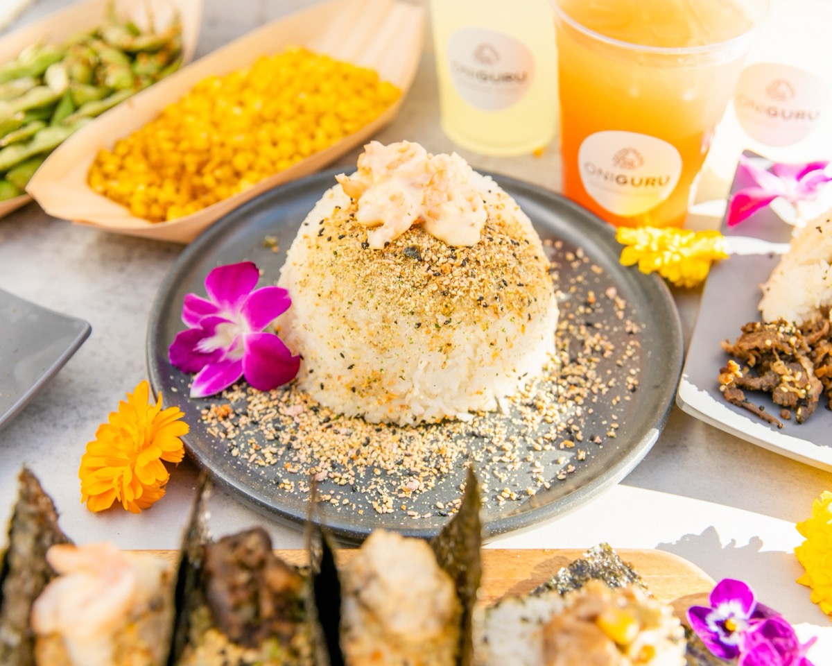 An OniGuru table setting: a hand-shaped rice bowl topped with crab on a grey plate, with nori-wrapped onigiri, edamame, corn and two OniGuru-branded drinks around it