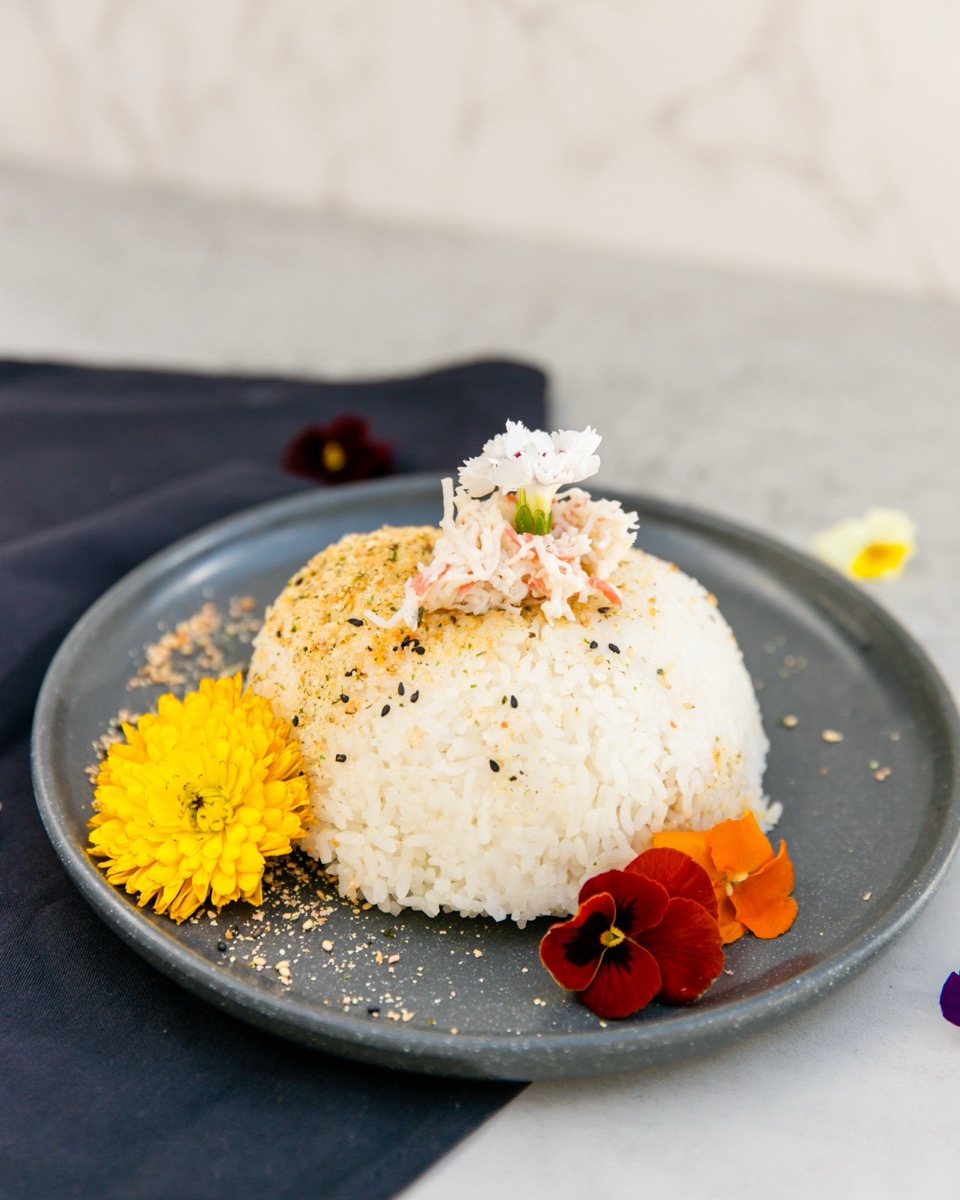 An OniGuru rice bowl on a grey plate: a hand-shaped dome of white rice dusted with furikake, topped with crab and an edible flower