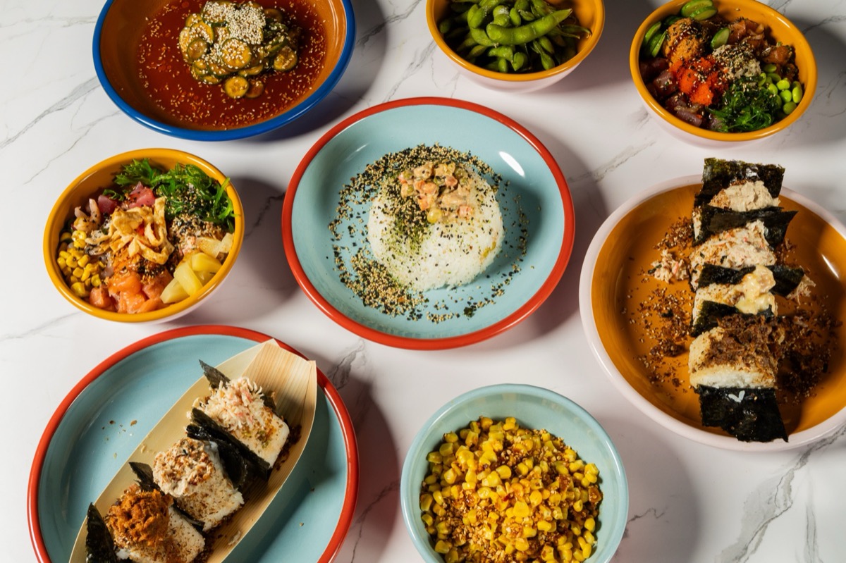 Overhead spread of OniGuru dishes: two poke bowls, a hand-shaped rice bowl dusted with furikake, sliced nori-wrapped onigiri, edamame, corn and cucumber salad